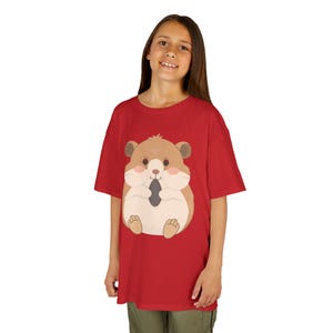 May include: A red t-shirt featuring a cartoon hamster design. The hamster is brown, beige, and gray, with pink cheeks and a dark gray object in its mouth. The shirt is worn by a person.