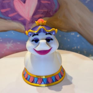 May include: A white figurine of a character with a purple hat and a gold crown. The figurine has blue eyes, a pink mouth, and a decorative base with blue, pink, and gold accents. The background features a heart shape.