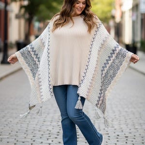 May include: A beige ribbed knit sweater layered under a cream-colored poncho featuring a multi-colored zigzag pattern in shades of grey, blue, and tan, finished with decorative tassels along the bottom hem, paired with blue denim jeans and white sneakers.