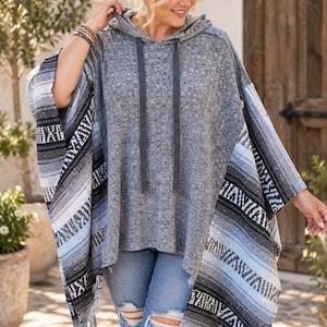 May include: A gray hooded poncho featuring a textured knit center panel and wide side panels with a horizontal striped pattern in shades of blue, black, white, and gray. The garment includes a drawstring hood and a fringed hem along the bottom edges.