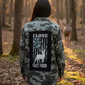 May include: A camouflage-patterned shirt with a black patch featuring a white deer silhouette and the text "I LOVE FAST FOOD" in white. The shirt is worn in a forest setting, with a deer in the background.