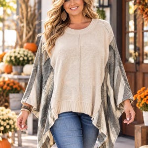 May include: A beige knit sweater featuring a round neckline and a ribbed hem, paired with blue denim jeans. The sweater has wide, poncho-style sleeves with a dark gray and cream diagonal striped pattern that contrasts with the solid beige center panel.