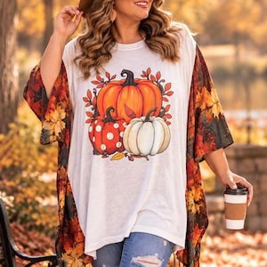 May include: A white t-shirt featuring a graphic of three pumpkins, one large orange, one red with white polka dots, and one white, surrounded by autumn leaves. The shirt is worn under a kimono-style cover-up with a vibrant fall leaf print pattern.