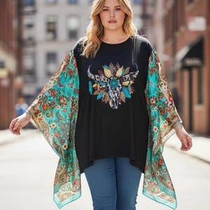 May include: A black tunic top featuring a central graphic of a bull skull adorned with colorful feathers and leopard print patterns. The garment has wide, flowing sleeves with a vibrant turquoise, orange, and brown paisley floral print design.