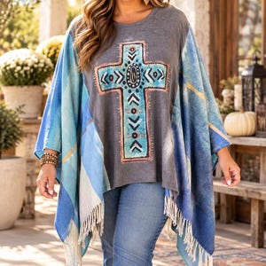 May include: A charcoal gray poncho-style top featuring a large, central turquoise and orange cross graphic with geometric patterns. The garment has wide, blue and white plaid-patterned sleeves and a hemline finished with long white fringe.