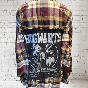 May include: A plaid flannel shirt in burgundy, gold, and cream, with a distressed Hogwarts patch on the back. The patch features the word "HOGWARTS" and the school's crest with the motto "Draco Dormiens Nunquam Titillandus."