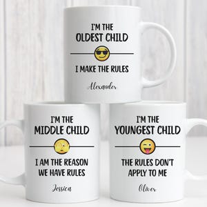 May include: Three white ceramic mugs with black text and emoji designs. The mugs read "I'M THE OLDEST CHILD," "I'M THE MIDDLE CHILD," and "I'M THE YOUNGEST CHILD." Each mug has a different emoji and a name below the text.