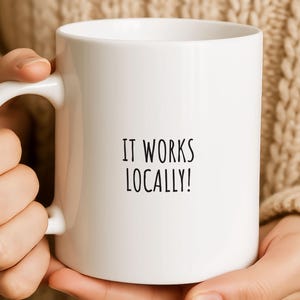 May include: White ceramic mug with a curved handle. The mug features the text "IT WORKS LOCALLY!" in black, centered on the front. The mug is held by a person wearing a beige sweater.