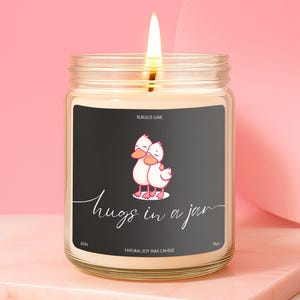 May include: A lit candle in a glass jar with a black label. The label features a cartoon illustration of two ducks hugging and the words "hugs in a jar". The candle is labeled "Natural Soy Wax Candle" and is on a pink surface.