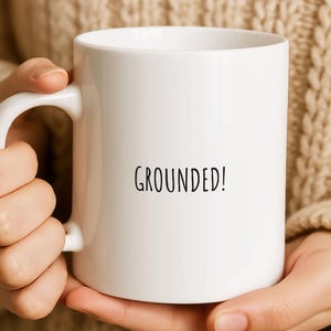 May include: White ceramic mug with the word "GROUNDED!" printed in black. The mug has a curved handle and is held by a person wearing a beige sweater. The mug is a standard size for coffee or tea.