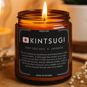 May include: A lit amber glass candle with a black label that reads "KINTSUGI" and "JAPANESE." The candle sits on a wooden tray with small white flowers. The text on the label describes the art of embracing imperfections.