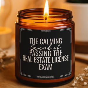 May include: A lit amber glass candle with a black label that reads "THE CALMING Scent of PASSING THE REAL ESTATE LICENSE EXAM." The candle is on a wooden tray with small white flowers. The text "NATURAL SOY WAX CANDLE" is at the bottom.