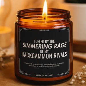 May include: A lit soy wax candle in an amber glass jar with a black label. The label reads: RUBULUS LUME, FUELED BY THE SIMMERING RAGE OF MY BACKGAMMON RIVALS, Infused with lucky doubles, smooth bearing off, and the ancient art of looking calm while winning.