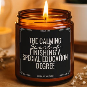 May include: A lit soy wax candle in a brown glass jar. The black label reads "THE CALMING Scent of FINISHING A SPECIAL EDUCATION DEGREE." The candle sits on a wooden tray, with a small plant and light in the background.