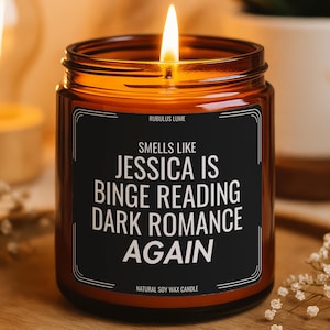 May include: A lit candle in an amber glass jar with a black label. The label features the text: RUBULUS LUME, SMELLS LIKE JESSICA IS BINGE READING DARK ROMANCE AGAIN, and NATURAL SOY WAX CANDLE. The candle has a single flame burning at the top.