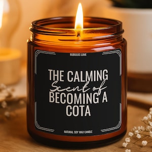 May include: A lit soy wax candle in a brown glass jar. The black label reads "THE CALMING Scent of BECOMING A COTA". The candle sits on a wooden tray with small white flowers.