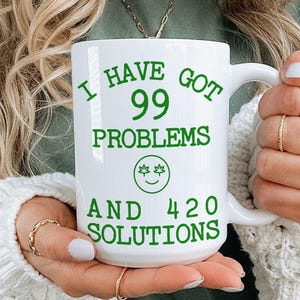 May include: White ceramic mug with the text "I HAVE GOT 99 PROBLEMS AND 420 SOLUTIONS" in green. The mug also features a green smiley face with two leaves. The mug is held by a person.