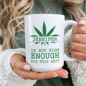 May include: White ceramic mug with a green cannabis leaf design and the name "Jennifer" printed above the phrase "IS NOT HIGH ENOUGH FOR THIS SHIT" in green. The mug is held by a person.
