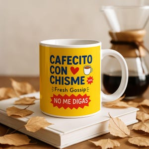 May include: A white ceramic coffee mug with a yellow background featuring the text "CAFECITO CON CHISME" and "Fresh Gossip". The mug also has a red heart and a coffee cup icon. The phrase "NO ME DIGAS" is in a red starburst.