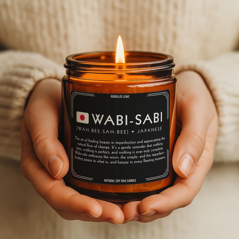 Może przedstawiać: Zapalone bursztynowe szklane naczynie na świece z czarną etykietą z napisem "WABI-SABI" białym tekstem. Świeca jest oznaczona jako "Natural Soy Wax Candle". Świeca jest trzymana w dłoniach osoby.