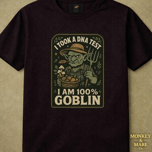 May include: A black short-sleeved t-shirt featuring a graphic of a green goblin wearing a straw hat, holding a pitchfork and a basket of mushrooms. The text reads I TOOK A DNA TEST I AM 100% GOBLIN. The design is framed in a rectangular border.