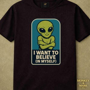 Könnte beinhalten: Ein schwarzes T-Shirt mit kurzen Ärmeln und einer Grafik eines grünen Aliens mit verschränkten Armen in einem blaugrünen rechteckigen Rahmen. Unter dem Alien steht der Text I WANT TO BELIEVE (IN MYSELF) in weißen Großbuchstaben auf dunkelblauem Grund.