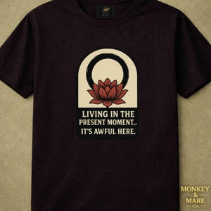 May include: A dark brown short-sleeved t-shirt featuring a graphic of a red lotus flower inside a black circle and an arch. Below the graphic, the text reads: LIVING IN THE PRESENT MOMENT... IT'S AWFUL HERE. The shirt has a crew neckline.