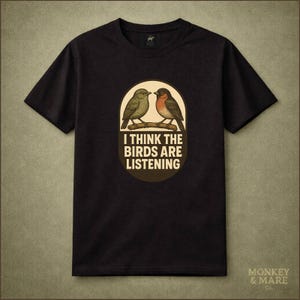 May include: Black t-shirt featuring a graphic of two birds perched on a branch inside an oval. The text "I THINK THE BIRDS ARE LISTENING" is below the birds. The shirt has short sleeves and a crew neck.