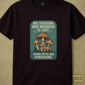 Puede incluir: Camiseta granate oscuro con un diseño rectangular que presenta setas y el texto "NOT EVERYONE WHO WANDERS IS LOST. SOME OF US ARE FORAGING." El diseño incluye un fondo verde azulado con estrellas y una base de hierba. El logotipo de la marca está en la esquina inferior derecha.
