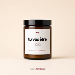 May include: A brown glass candle jar with a black lid and a white label. The label features red quotation marks, the black text "tu vas être tata," and the red text "bougie artisanale • pamplemousse." The brand name "maison flambeuse" appears below.