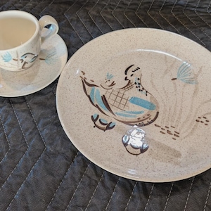 Puede incluir: Un juego de taza y platillo de té de cerámica vintage con un plato llano a juego. El juego presenta un fondo beige moteado con un diseño de pájaro y floral en marrón y turquesa. La taza y el platillo están sobre una superficie acolchada gris oscuro.