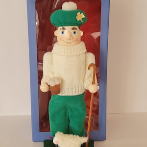 May include: A wooden nutcracker figurine of a man in a green hat with a shamrock, white sweater, and green pants, holding a mug and a cane. A small sheep stands on a green base with an Irish flag.