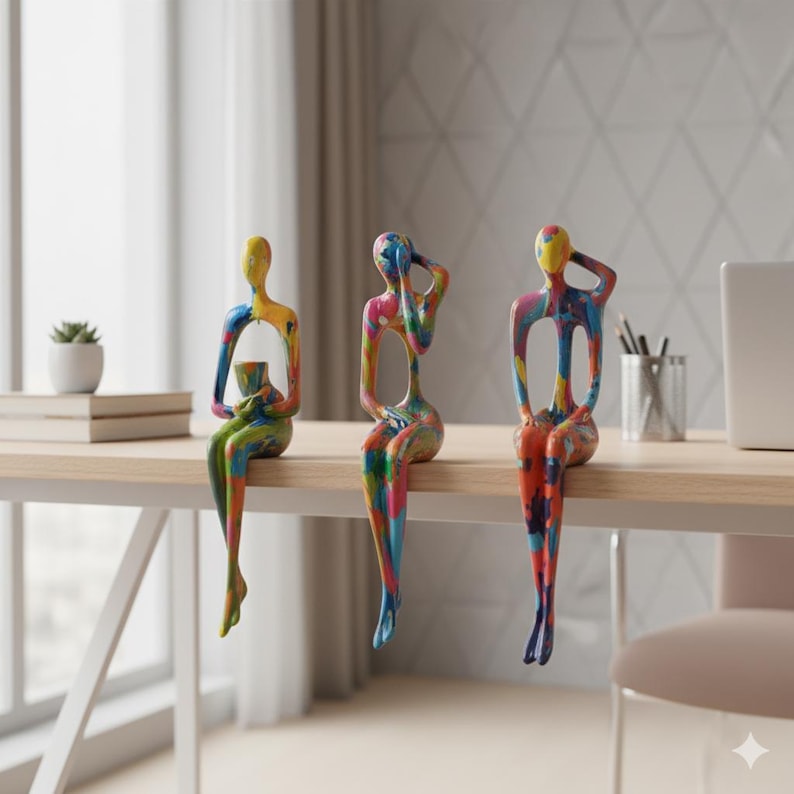 May include: Three colourful abstract sculptures of seated figures with arms raised, painted in a vibrant mix of colours. The sculptures are placed on a light-coloured wooden desk, with a plant, books, and office supplies in the background. The figures are approximately 25 cm tall.