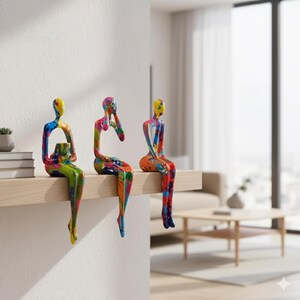 May include: Three abstract sculptures of seated figures with colourful paint splatter designs. Each figure is perched on a wooden shelf, showcasing a modern art aesthetic. The sculptures feature yellow, blue, green, and red hues.