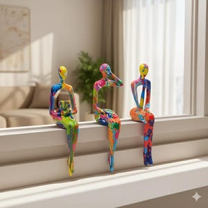 May include: Three abstract sculptures of seated figures with colourful paint splatter designs. The figures are perched on a white windowsill. The sculptures are in shades of yellow, blue, green, pink, and orange.