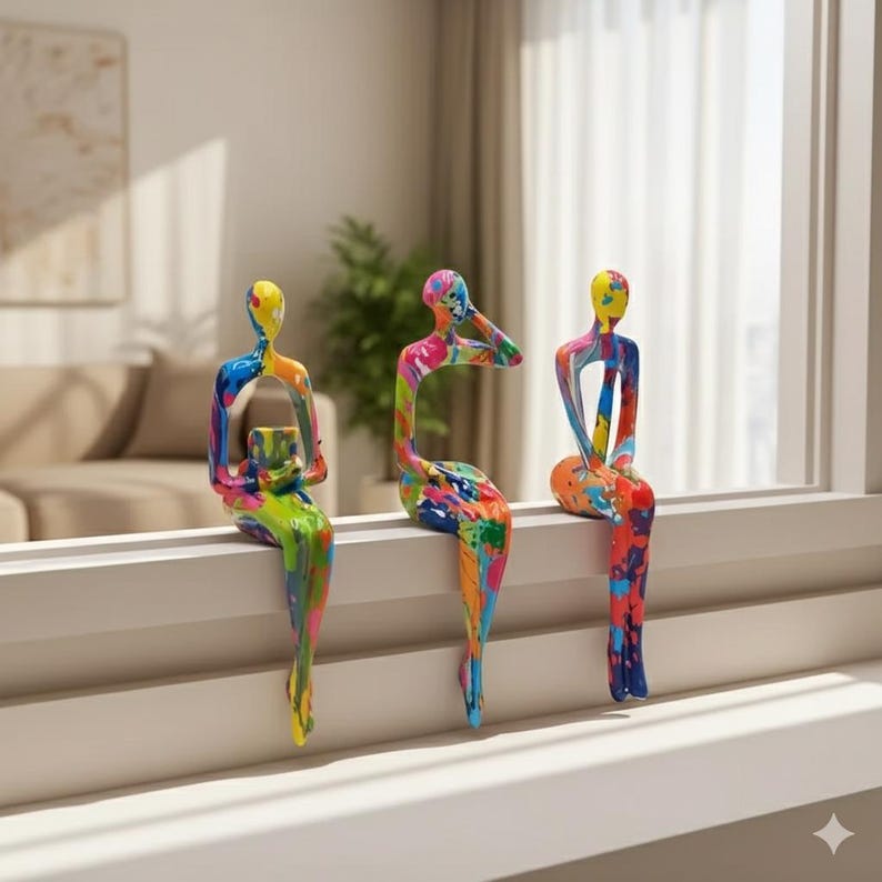 May include: Three abstract sculptures of seated figures with colourful paint splatter designs. The figures are perched on a white windowsill. The sculptures are in shades of yellow, blue, green, pink, and orange.