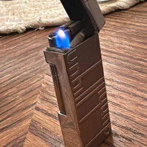 May include: A rectangular, brown and silver lighter with a blue flame. The lighter is open, revealing the flame. The lighter has a decorative design on the side and is on a wooden surface.