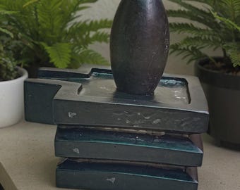 May include: A dark, oval-shaped stone sits atop a dark green, layered, rectangular base. The stone has a smooth, polished surface. The base has a textured appearance. Green plants are visible in the background.