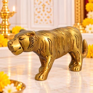 May include: A gold-coloured metal tiger figurine standing in a profile position. The sculpture features engraved stripes along its body, a detailed face with visible ears, and a tail curved upwards. The surface has a polished, metallic finish.