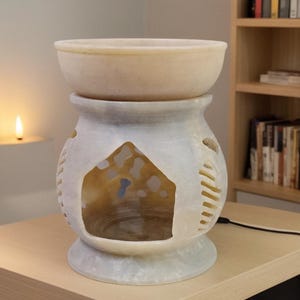 Op de afbeelding: Een witte en beige essentiële olie diffuser met een komvormige bovenkant en een decoratieve basis. De basis heeft uitgesneden ontwerpen en een huisvormige opening. De diffuser is gemaakt van een steenachtig materiaal.