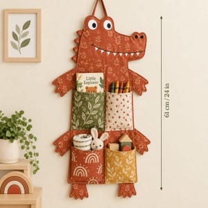 May include: A wall-hanging fabric organizer shaped like a crocodile in rust orange with a leaf pattern. It features four pockets containing a book titled Little Explorer, crayons, a small plush rabbit, and wooden blocks. It measures 24 inches in height.
