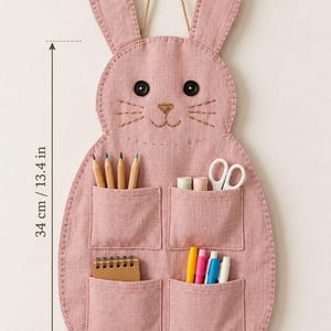 Puede incluir: Un organizador de pared de tela rosa empolvado con forma de conejo y cuatro bolsillos que contienen lápices, tijeras y rotuladores. El organizador tiene ojos de botón negros, bigotes bordados y un cordón para colgar. Mide 34 cm de altura.