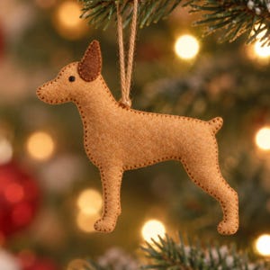May include: A tan felt dog ornament with a dark brown ear and a small black eye. The dog is stitched along the edges with tan thread and hangs from a thin, light brown jute string loop, designed for display as a decorative hanging item.