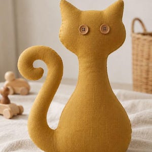 Könnte beinhalten: Eine senfgelbe Stoffkatzenfigur mit einem geschwungenen Schwanz und zwei runden, hellbraunen Knöpfen als Augen. Die Katze hat eine einfache, genähte Silhouette und ist aus einem strukturierten Textilmaterial gefertigt, aufrecht stehend.