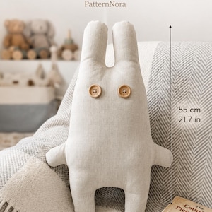 Puede incluir: Un juguete de peluche de tela beige con dos orejas largas y dos botones de madera redondos como ojos. El juguete mide 55 cm de alto y 39 cm de ancho. El texto PDF PATTERN y PatternNora es visible en la parte superior.