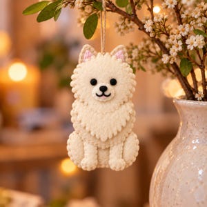 May include: A cream-colored felt dog ornament shaped like a Pomeranian with a fluffy layered chest, small pink-lined ears, black bead eyes, and a stitched black nose and mouth, suspended by a thin twine loop for hanging as a decorative piece.