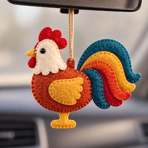 May include: A felt rooster hanging ornament featuring a red comb, cream-colored face, brown body, and a yellow wing. The tail consists of layered felt feathers in blue, yellow, and red. It hangs from a tan string attached to a loop at the top.