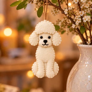 May include: A cream-colored felt poodle ornament featuring a stitched edge, black bead eyes, and a black stitched nose and mouth. The dog figure is suspended by a thin brown twine loop, showcasing a detailed silhouette with a curly head design.