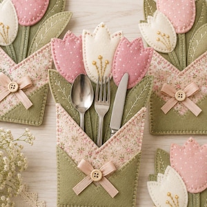 May include: A set of felt cutlery holders shaped like tulip bouquets. Each holder features pink and white felt tulips, green felt leaves, and a floral fabric pocket accented with a small beige ribbon and a wooden button. A silver spoon, fork, and knife are tucked inside the central holder.