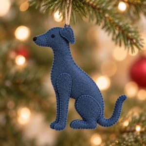 May include: A dark blue felt dog ornament featuring visible stitched edges and a small black bead eye. The dog is in a seated position with a curved tail and is suspended by a thin tan twine loop attached to the top of its head for hanging.